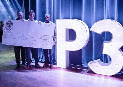 Three men hold a large donation check and stand next to a lighted marquee sign