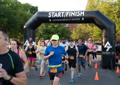 A man in a blue shirt and neon yellow Nike hat runs past the Start/Finish banner among a crowd of runners.