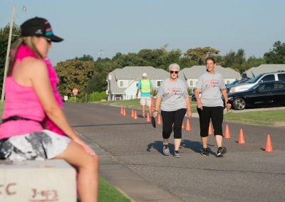 Two women wearing black capris and matching Pink Up the Pace t-shirts walk past a woman sitting on the sidelines.