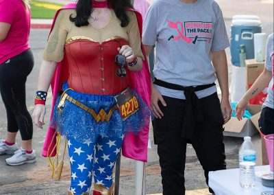 A woman dressed as Wonder Woman smiles next to a breast cancer survivor at a breast cancer awareness race.