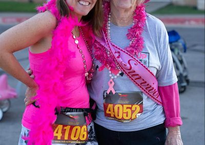 A younger woman wearing a pink feather boa standing next to an older woman wearing a pink hat and a pink sash that says "Survivor"