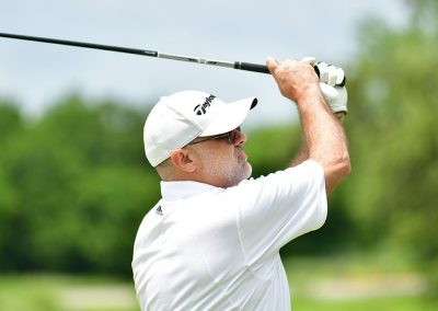 A man wearing a white hat and white golf polo swings a golf club