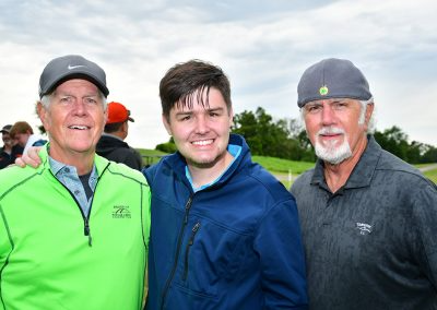 A young man with brown hair stands in between two older man wearing gray hats at a breast cancer awareness golf tournament.