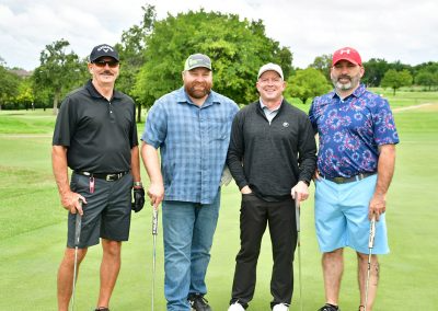 Four middle-aged men smile for a photo while holding their golf clubs on a golf course.