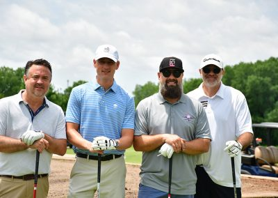 Four middle-aged men hold their golf clubs and stand on a golf course.