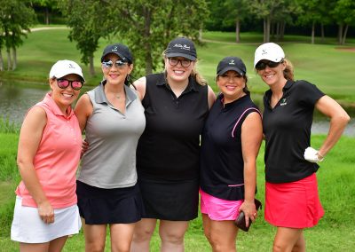 A group of five women wearing golfing attire and hats smile for a photo on a golf course.