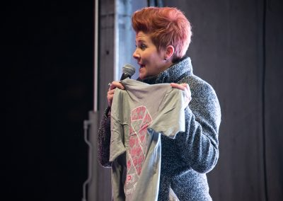 A breast cancer survivor holding up shirt and speaking into a microphone at a breast cancer awareness event