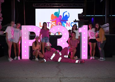 A group of teenage girls gather around a P31 marquee sign as a man wearing a pink firefighter uniform lays on the ground in front