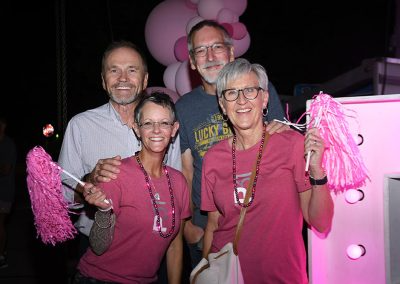 Two older women wearing pink shirts and holding pink pom poms crouch in front of their husbands