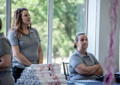 A woman with a ponytail crosses her arms and listens while sitting next to a woman that is standing