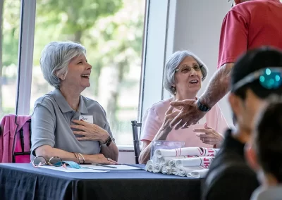 Two older women laugh while sitting behind a raffle table at a breast cancer golf tournament