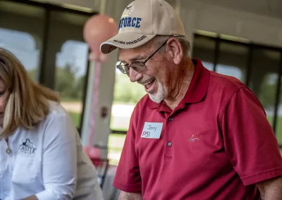 An older man wearing a tan Air Force hat and red golf polo smiles as he looks down at a table