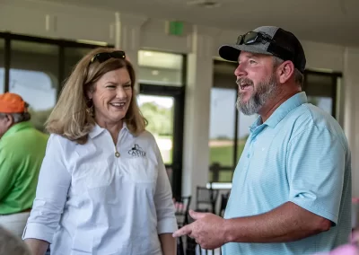 A middle-aged man wearing a light blue golf polo talks to an older woman in a white shirt