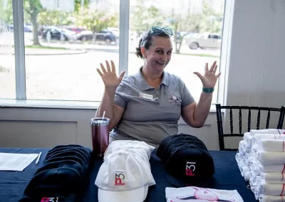 An older woman smiles and raises both hands while sitting behind a table full of hats