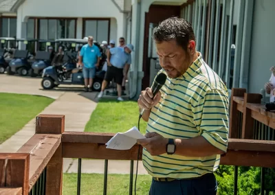 A middle-aged man wearing a striped polo reads a paper while speaking to a crowd at a breast cancer golf event