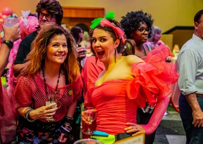 two women wearing neon clothing and holding wine glasses pose for a photo at a breast cancer awareness event