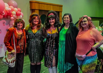 Women wearing bright and sparkly costumes smile and pose for a group photo next to a photobooth