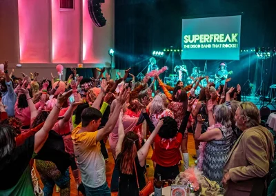 A crowd of people wearing costumes raise their hands in the air as the band plays on stage