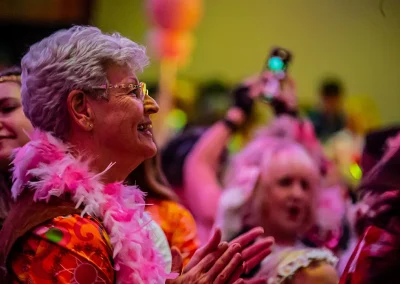 An older woman wearing a pink feather boa claps and smiles