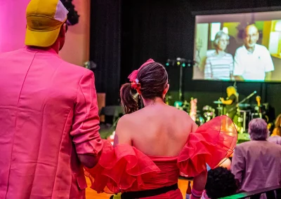 A man and woman wearing bright and colorful customes watch as a video plays on a large screen