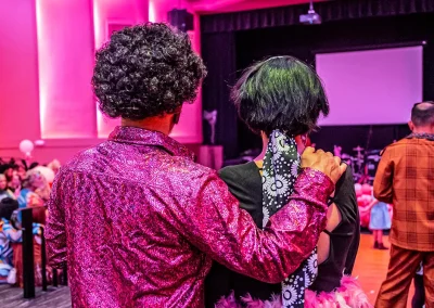 A man and a woman wearing 70s costumes hug while looking at dancefloor