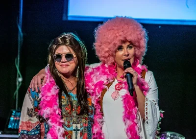 A woman wearing a pink afro wig and talking into a microphone hugs another woman wearing sunglasses and a pink feather boa.