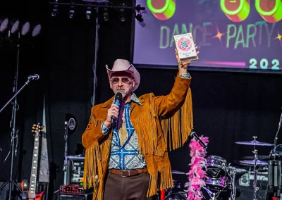 Man wearing a fringe jacket and cowboy hat holding up a small sign while speaking into a microphone.