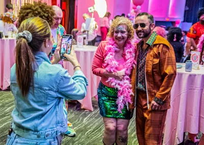 Man and woman wearing 70s costumes smiling as young woman takes their picture on cell phone.