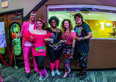 Group of adults wearing colorful neon 80s costumes and holding appetizers