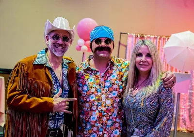 Three middle-aged adults wearing retro costumes smile for a photo at a breast cancer awareness gala