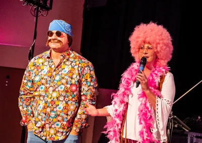A breast cancer survivor wearing a big afro wig and a pink feather boa talks into a microphone on stage with a man wearing a retro outfit next to her