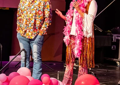 A middle-aged man wearing a retro costume stands onstage next to a middle-aged woman wearing a pink afro wig.