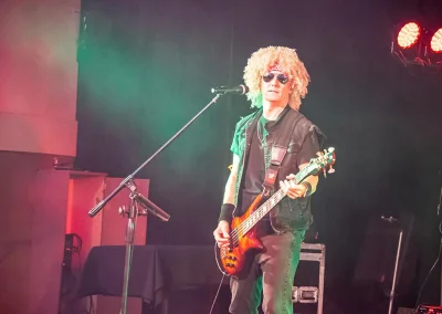 A middle-aged man wearing a fake afro wig and sunglasses plays guitar on stage