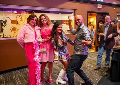 Two women wearing bright pink dresses smile as a middle-aged man and woman wearing 70s costumes dance