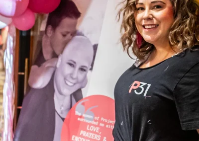 A younger woman with curly hair smiles and stands next to a poster for breast cancer awareness