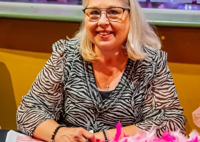 An older woman with grey hair and glasses smiles with a pen and spreadsheet in her hands