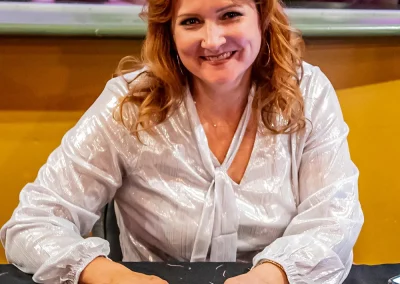 A middle-aged woman with red hair smiles with a pen and spreadsheet