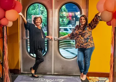 Two women pose with one arm raised while standing in front of a set of silver doors