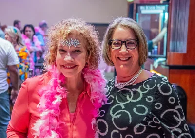 An older woman with curly hair and jewels attached to her forehead smiles with another woman wearing glasses