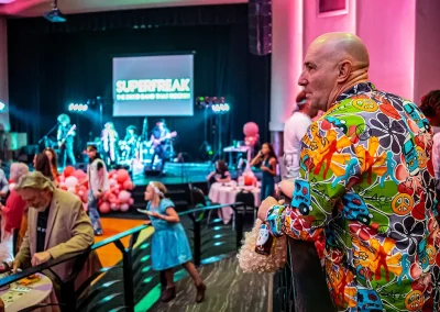 A middle-aged man wearing a bright suit overlooks the dance floor while holding a glass bottle