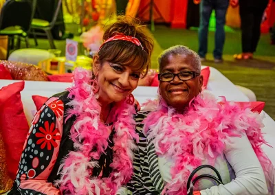 Two older women wearing pink feather boas smile while sitting on a white couch