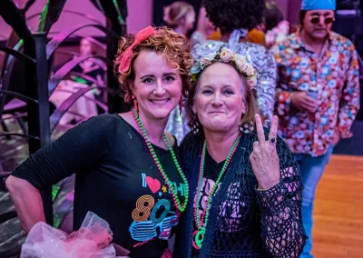 An older woman wearing a 70s costume holds up a peace sign as she poses for a photo with a younger woman