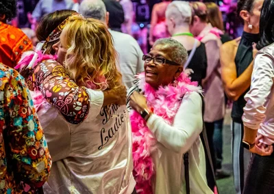 An older woman with glasses and a feather boa smiles at two middle-aged women hugging