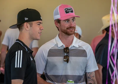 A young man with red hair wearing a pink ball cap stands and talks with a teenage boy wearing a black ball cap.