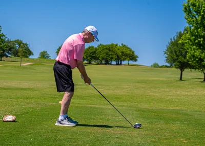 An older man wearing a pink polo gets ready to hit a golf ball with a driver at the Sink4Pink Golf Tournament.