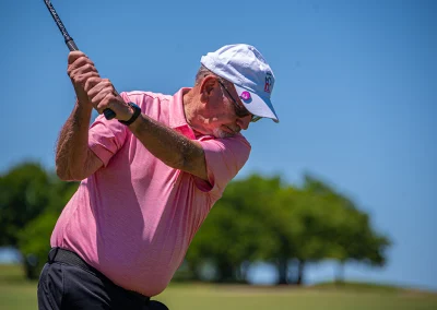 An older man wearing a pink polo and a Project31 ball cap swings to hit a golf ball.