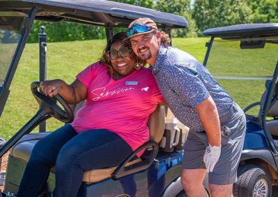 A woman with a bright pink shirt and large sunglasses hugs a male golfer while sitting in a parked golf cart.