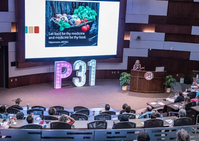 A breast cancer survivor speaks to a large crowd at the 2023 survivorship expo. She is on a stage with a P31 marquee and a slideshow.