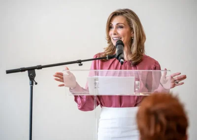 A woman in a pink shirt and white slacks gives a speech at the podium.