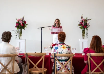 A woman in a pink shirt and white slacks gives a speech at the podium.